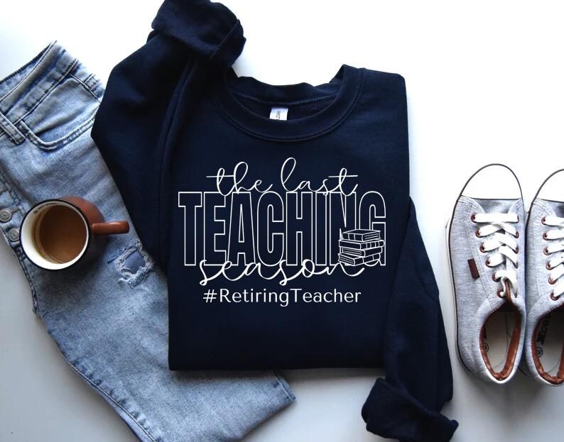 K&ouml;nnte beinhalten: Ein marineblauer Sweatshirt mit dem Text "The Last Teaching Season #RetiringTeacher" in Wei&szlig;. Der Text ist von einem Stapel B&uuml;cher umgeben.