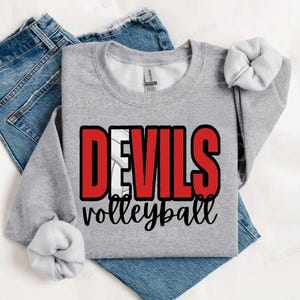 Puede incluir: Una sudadera gris con la palabra "DEVILS" en letras rojas y un gráfico de voleibol blanco. Debajo de la palabra "DEVILS" está la palabra "volleyball" en cursiva. La sudadera está colocada sobre unos vaqueros azules.