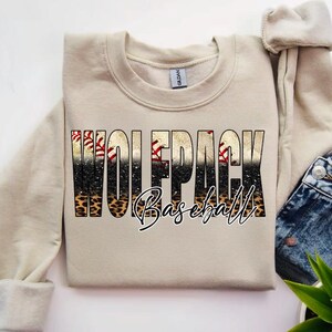 以下が含まれることがあります： 「WOLFPACK Baseball」の文字が入ったベージュのスウェットシャツ。文字は野球とヒョウ柄のデザイン。スウェットシャツはデニムジーンズと緑の植物と組み合わせています。カジュアルなアイテムです。