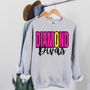 May include: Gray sweatshirt with the text "DIAMOND Divas" in pink and black letters. A yellow softball with black stitching is in the middle of the word "DIAMOND".