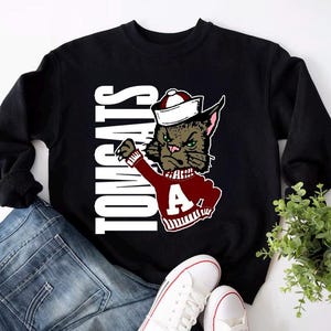 May include: Black sweatshirt with the word "TOMCATS" in white vertical text. The sweatshirt features a cartoon cat wearing a sailor hat and a maroon sweater with a white "A". The sweatshirt is paired with blue jeans and white sneakers.