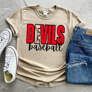 Puede incluir: Una camiseta beige con el texto "DEVILS baseball" en letras rojas y negras. La palabra "DEVILS" tiene un gráfico de béisbol detrás.