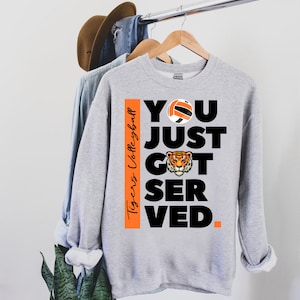 Puede incluir: Sudadera gris con un gr&aacute;fico negro y naranja que dice "You Just Got Served." El gr&aacute;fico tambi&eacute;n incluye un bal&oacute;n de voleibol y una cabeza de tigre. El texto "Tigers Volleyball" est&aacute; en el lado del gr&aacute;fico.
