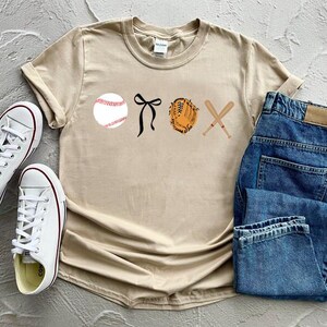 May include: A beige t-shirt with a baseball, a glove, bats, and a bow printed on the front. The shirt is laying on a surface with a pair of white sneakers and a pair of blue jeans.