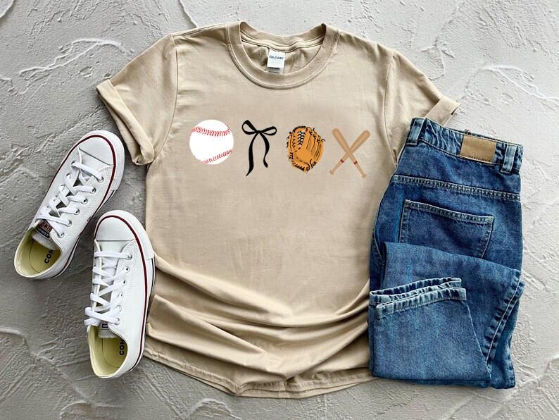 May include: A beige t-shirt with a baseball, a glove, bats, and a bow printed on the front. The shirt is laying on a surface with a pair of white sneakers and a pair of blue jeans.