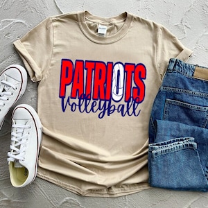 Peut inclure: Un t-shirt beige avec le texte "Patriots Volleyball" en lettres rouges, bleues et blanches. Le t-shirt est pli&eacute; et pos&eacute; sur une surface blanche avec une paire de baskets blanches et un jean bleu.