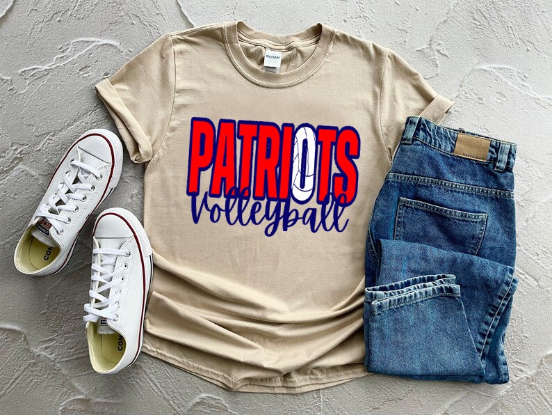 Peut inclure: Un t-shirt beige avec le texte "Patriots Volleyball" en lettres rouges, bleues et blanches. Le t-shirt est pli&eacute; et pos&eacute; sur une surface blanche avec une paire de baskets blanches et un jean bleu.