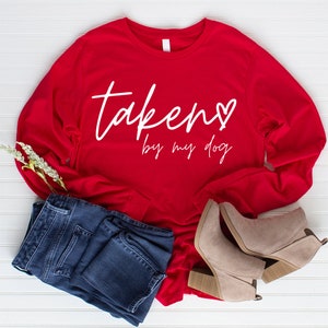 May include: A red long-sleeve shirt with the text "taken by my dog" in white script. The shirt is paired with blue jeans and beige ankle boots. A sprig of white flowers is also present.