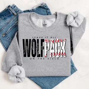Könnte beinhalten: Grauer Pullover mit dem Aufdruck "Leave it all Wolfpack Baseball on the field" in Schwarz. Das Wort "pack" ist teilweise von einem Baseball-Motiv mit roten Nähten verdeckt.