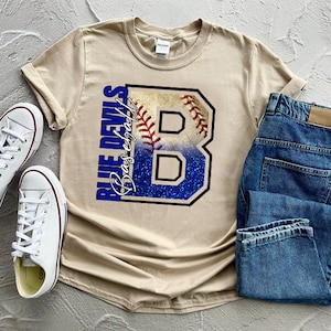 May include: A tan t-shirt with a blue and white baseball graphic and the text "Blue Devils Baseball" printed on the front. The shirt is paired with blue jeans and white sneakers.
