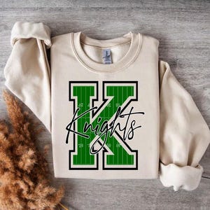May include: A cream-coloured sweatshirt with a green and black letter "K" with the word "Knights" written in black script. The letter "K" is designed to look like a football field.