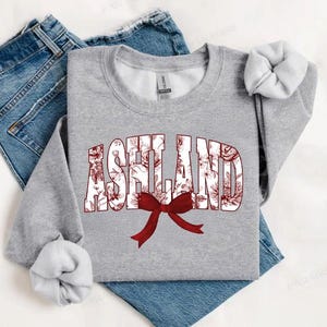 May include: A gray sweatshirt with the word "ASHLAND" printed in white with a red floral pattern. A red bow is centered below the word.