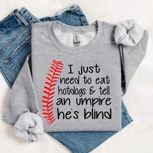 Op de afbeelding: Een grijs sweatshirt met een rood baseball-steekontwerp en de tekst "I just need to eat hotdogs & tell an umpire he's blind".
