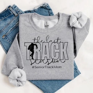 May include: Gray sweatshirt with black text that reads "the last track season #SeniorTrackMom". A silhouette of a runner is in the word "track".