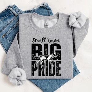 May include: A heather gray sweatshirt with the text "Small Town Big Pride" in black. A black and white dog graphic is in the center. The sweatshirt is folded on top of a pair of blue jeans.
