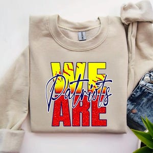 May include: A beige sweatshirt with the text "We Are Patriots" in red, yellow, and blue. The text is layered over a softball graphic.