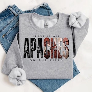 May include: A heather gray sweatshirt with the words "LEAVE IT ALL APACHES FOOTBALL ON THE FIELD" in black and red. The word "FOOTBALL" is designed with a football texture. The sweatshirt is laid on top of blue jeans.