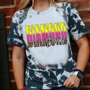 May include: A white t-shirt with a tie-dye pattern on the sleeves and shoulders. The shirt features the words "DIAMOND Divas" in a colorful design with a leopard print accent. The shirt is a casual wear item.