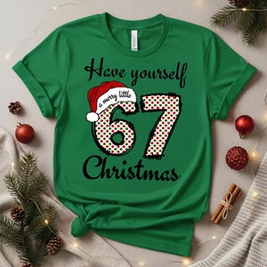 Op de afbeelding: Groen t-shirt met de tekst "Have yourself a merry little 67 Christmas." Het getal 67 is versierd met een rood-groen stippenpatroon en een kerstmuts. De tekst is zwart. Kerstkleding.
