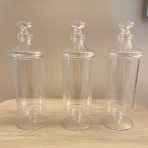 May include: Three clear plastic jars with lids and pedestal bases. The jars are empty and are arranged in a row on a light wood surface.