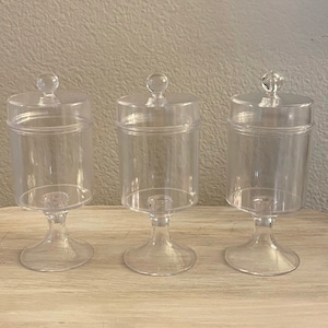 Puede incluir: Tres soportes para tartas de pedestal de plástico transparente con tapas. Los soportes están sobre una superficie de madera clara.