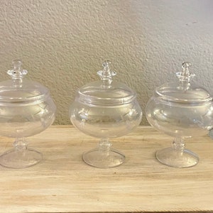 May include: Three clear glass candy jars with lids on a wooden surface. The jars have a pedestal base and are decorated with a subtle etched pattern.
