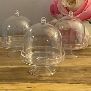May include: Three clear plastic cake stands with dome lids. The stands are on a wooden surface.