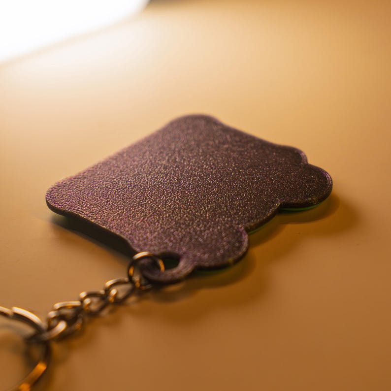 May include: A dark purple keychain with a textured, glittery surface and a rounded, scalloped edge shape. The keychain features a small circular hole at the bottom, which is connected to a metallic chain and a circular split ring for attachment.