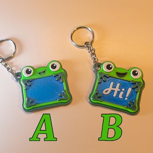 May include: Two frog-shaped keychains with metal rings. Keychain A features a solid blue center, while keychain B features a blue center with the white text Hi! printed on it. Both keychains have green borders, white eyes with black pupils, and small frog feet.