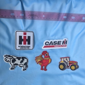 May include: A collection of five embroidered patches including a square International Harvester logo, a Case IH Agriculture logo, a black and white cow, a red rooster, and a red tractor, positioned below a ruler measuring 12 inches in length.