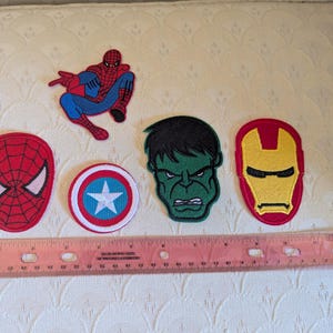May include: A collection of five embroidered superhero patches featuring Spider-Man, a Captain America shield, the Hulk, and Iron Man, positioned above a pink 12-inch ruler that shows the scale of the items in inches and centimeters.