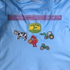 May include: A light blue fabric background featuring five embroidered patches: a black and white cow, a red rooster, two green tractors, and a yellow John Deere Quality Farm Equipment logo. A 12-inch purple ruler is positioned horizontally above the patches.