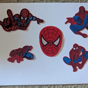 May include: Five embroidered fabric patches featuring the character Spider-Man in red and blue, including four full-body poses and one mask emblem. A wooden ruler is placed on the left side, showing the patches range from 2 to 5 inches in height.