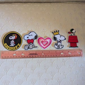 May include: A set of five embroidered patches featuring Snoopy characters and a heart, arranged horizontally above a 12-inch plastic ruler. The designs include a circular Snoopy patch, a heart-holding Snoopy, a pink heart, a crowned Snoopy, and Snoopy on a red doghouse.