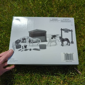 May include: A boxed toy set featuring miniature farm animals and accessories. The set includes a stable, horses, a wheelbarrow, and other farm-related items. The box is white with a warning label and a barcode.