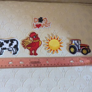 May include: A set of embroidered patches featuring a black and white cow, a red rooster, a yellow sun, a red tractor, and a white patch with the text "DOG LOVER" with a paw print and heart. A 12-inch ruler is placed below the patches for scale.
