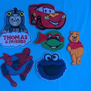 May include: A collection of six embroidered character patches featuring Thomas the Tank Engine with the text Thomas & Friends, a red race car, a green ninja turtle, a yellow bear in a red shirt, a red and blue superhero, and a blue monster face.