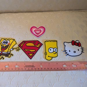 May include: A collection of five embroidered patches including a pink heart, a yellow Spongebob Squarepants, a red and yellow Superman logo, a yellow Bart Simpson head, and a white Hello Kitty face, positioned above a 12-inch wooden ruler.