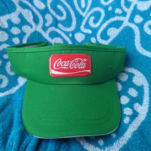 May include: A bright green visor featuring a rectangular red patch on the front panel. The patch displays the white Coca-Cola logo with a white swoosh underneath. The visor has a curved brim with visible stitching and an open top design.