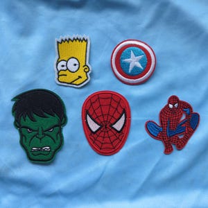May include: A collection of five embroidered fabric patches featuring a yellow cartoon character head, a red and blue circular shield with a white star, a green character head, a red spider-web patterned mask, and a red and blue spider-like figure.