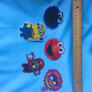 May include: Five embroidered character patches including a yellow minion, a blue monster, a red monster, a video game character in a red hat, and a purple creature, displayed next to a wooden ruler measuring approximately 12 inches in length.