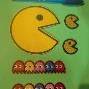 May include: A set of embroidered iron-on patches featuring the classic Pac-Man video game characters. The set includes a large Pac-Man, two smaller ghosts, and two rows of five ghosts in different colors.