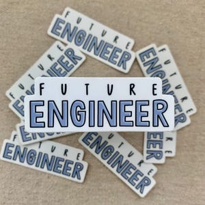 Puede incluir: Pegatinas rectangulares blancas con el texto "FUTURE ENGINEER" en fuente azul. Las pegatinas están esparcidas sobre una superficie beige. Un artículo divertido para estudiantes.