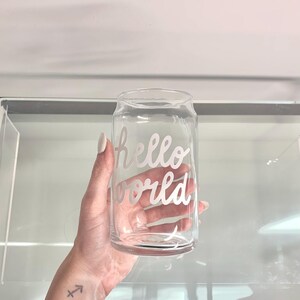 Può includere: Bicchiere trasparente a forma di lattina con le parole "hello world" in corsivo. Il bicchiere è tenuto da una persona. Il bicchiere è vuoto.