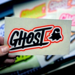 Könnte beinhalten: Ein weißer Aufkleber mit schwarzem und rotem Rand, auf dem das Wort "Ghost" in einer stilisierten Schriftart mit einer schwarzen Geister-Silhouette rechts neben dem Wort steht.