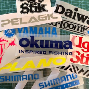 Puede incluir: Una colección de calcomanías de vinilo de marcas de pesca que incluyen logotipos de Okuma, Shimano, Daiwa, Plano, Pelagic y Ugly Stik en varios colores como azul, amarillo, rojo, negro y blanco, junto a un cúter sobre una base de corte verde.