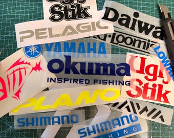 Puede incluir: Una colección de calcomanías de vinilo de marcas de pesca que incluyen logotipos de Okuma, Shimano, Daiwa, Plano, Pelagic y Ugly Stik en varios colores como azul, amarillo, rojo, negro y blanco, junto a un cúter sobre una base de corte verde.