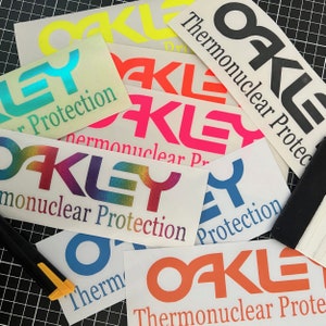May include: An assortment of colourful stickers with the word "Oakley" and the phrase "Thermonuclear Protection" on them. The stickers are on a black background.