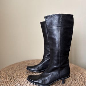 Puede incluir: Un par de botas negras de cuero hasta la rodilla con punta cuadrada y tacón pequeño. Las botas están sobre una superficie circular tejida. El cuero parece liso y las botas están en buen estado.