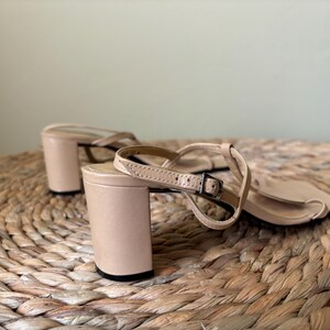 May include: A pair of beige sandals with a block heel and a thin ankle strap featuring a small metal buckle. The sandals have a square toe design and a smooth finish, with the heel and straps constructed in a matching light tan color.